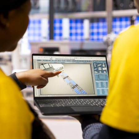 Smart factory experts use digital interface in optimization of industrial automation. Technicians review robotic assembly line and conveyor belt on laptop, enhancing efficiency in manufacturing plant.